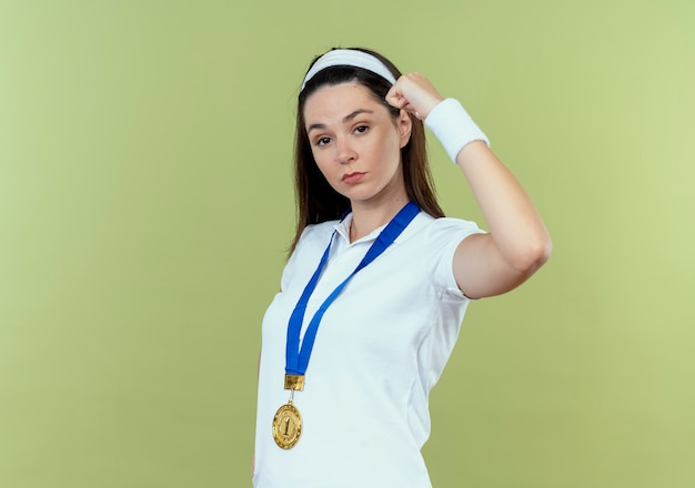 Молодая фитнес-женщина в повязке на голову с золотой медалью на шее, поднимая кулак, выглядит уверенно, стоя на светлом фоне