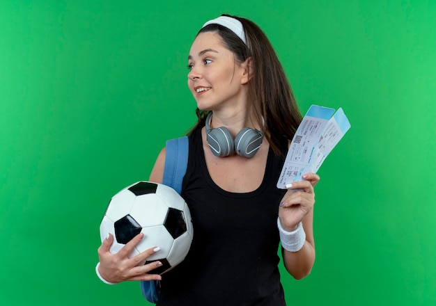 Молодая фитнес-женщина в головной повязке с наушниками и рюкзаком держит футбольный мяч и авиабилеты, глядя в сторону, улыбаясь, стоя на зеленом фоне