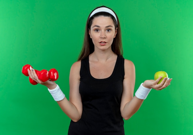 Young fitness woman in headband with headphones holding dumbbells and green apple looking at camera confused standing over green background