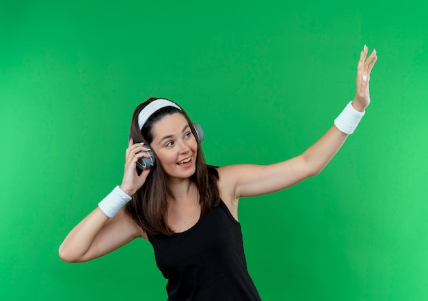 Giovane donna fitness in fascia con le cuffie che osserva da parte sorridendo allegramente agitando con una mano in piedi sopra il muro verde