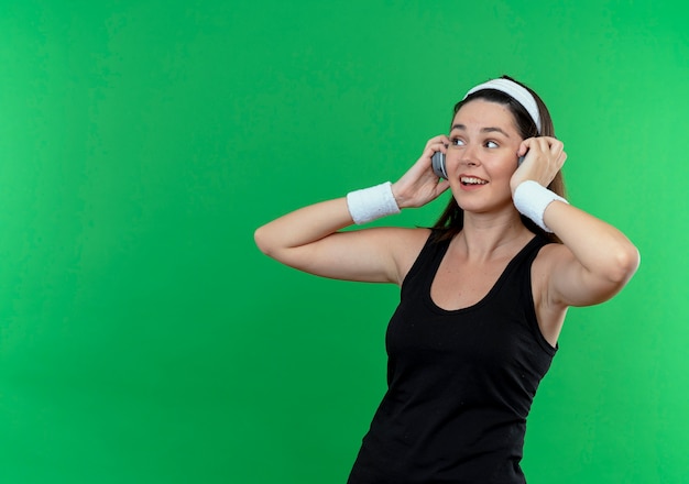 young fitness woman in headband with headphones looking aside smiling enjoying her favorite music standing over green wall