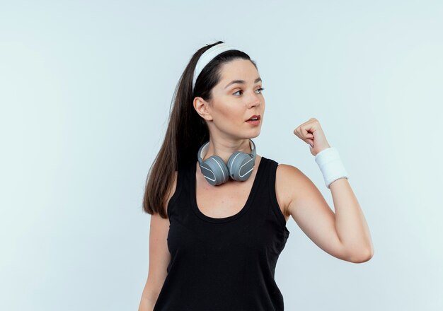 young fitness woman in headband with headphones looking aside with clenched fist standing over white wall