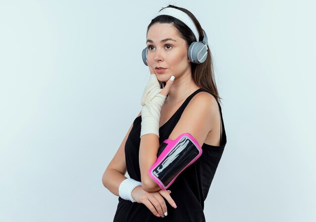 young fitness woman in headband with headphones and smartphone armband looking aside with pensive expression with hand on chin thinking standing over white wall