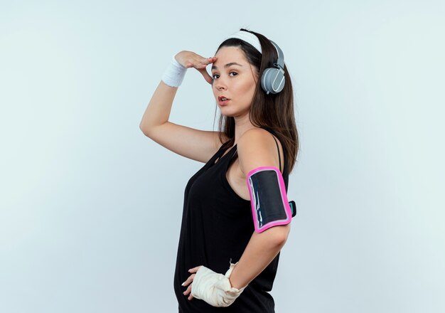 young fitness woman in headband with headphones and smartphone armband looking far away with hand over head standing over white wall