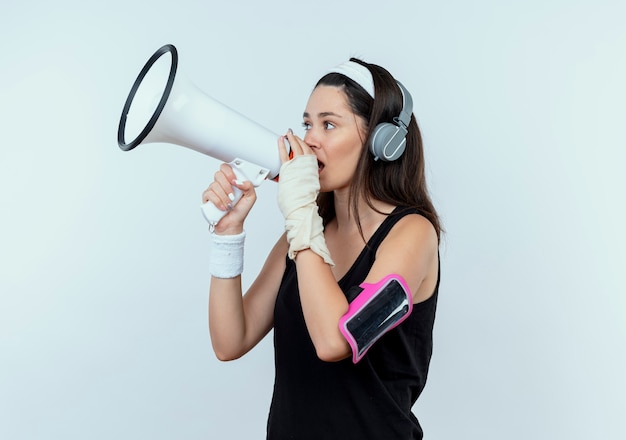 Молодая женщина фитнеса в повязке на голову с наушниками и повязкой смартфона кричит в мегафон, стоя на белом фоне
