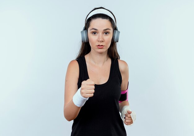 Молодая женщина фитнеса в повязке на голову с наушниками и повязкой на руке смартфона работает, глядя в камеру с серьезным лицом, стоящим на белом фоне
