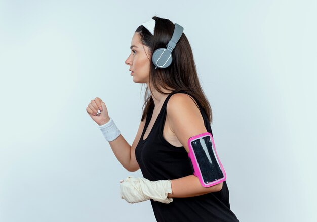 Молодая женщина фитнеса в повязке на голову с наушниками и повязкой на руке смартфона, тренирующейся стоя на белом фоне