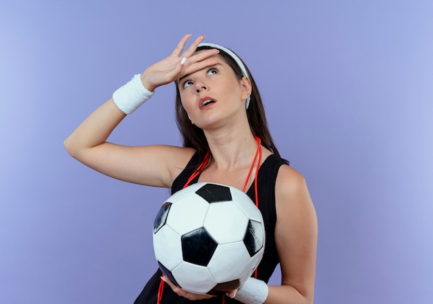 Young fitness woman in headband with skipping rope around neck holding soccer ball looking up tired standing over blue wall
