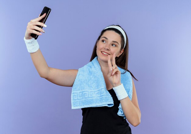 Young fitness woman in headband with towel around neck looking at screen of her smartphone taking selfie showing victory sing smiling standing over blue background