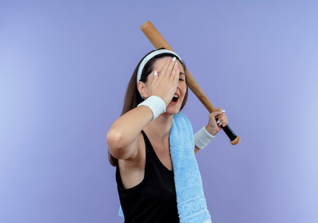 Молодая фитнес-женщина в повязке на голову с полотенцем на плече, держащая бейсбольную биту, прикрывая один глаз рукой, улыбаясь, стоя на синем фоне