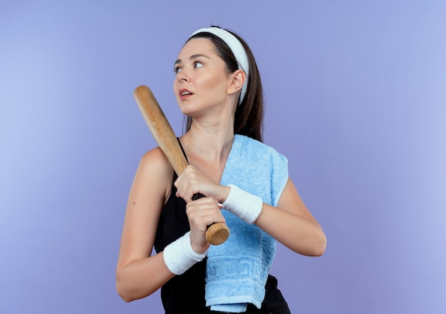 Young fitness woman in headband with towel on her shoulder holding baseball bat looking aside with serious face standing over blue background