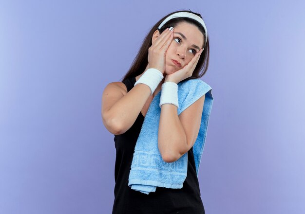 Young fitness woman in headband with towel on her shoulder looking aside bothered blowing cheeks standing over blue background