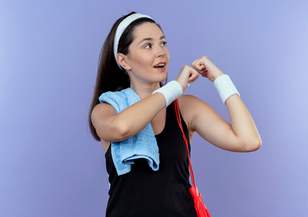 Young fitness woman in headband with towel on her shoulder looking aside clenching fist with confident expression standing over blue background