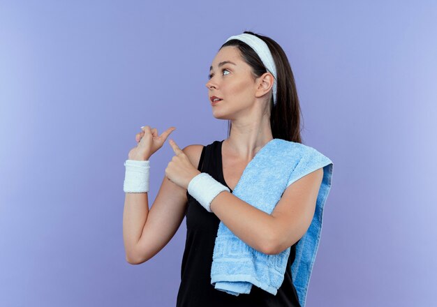 Young fitness woman in headband with towel on her shoulder looking aside pointing back standing over blue background