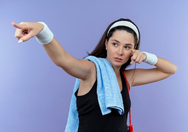 Young fitness woman in headband with towel on her shoulder looking confident pointing with fingers and hands to the side standing over blue wall
