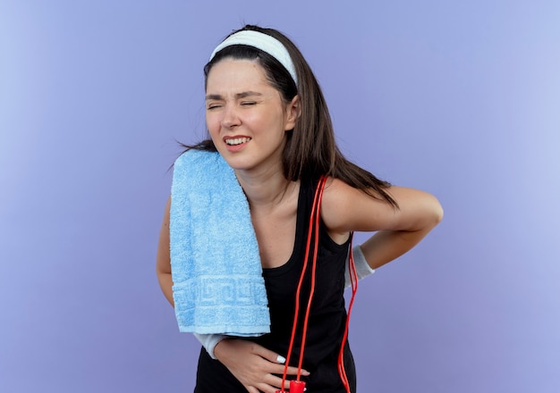 Giovane donna fitness in fascia con un asciugamano sulla sua spalla alla ricerca di malessere toccando la schiena sensazione di dolore in piedi su sfondo blu
