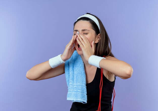 Young fitness woman in headband with towel on her shoulder shouting or calling someone with hands near mouth standing over blue background