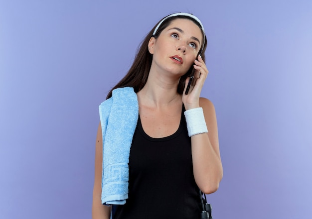 Young fitness woman in headband with towel on her shoulder talking on mobile phone looking up with pensive expression standing over blue wall