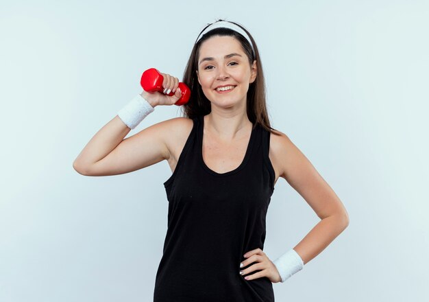Молодая женщина фитнеса в оголовье, тренирующемся с гантелями, глядя в камеру, улыбаясь, стоя на белом фоне
