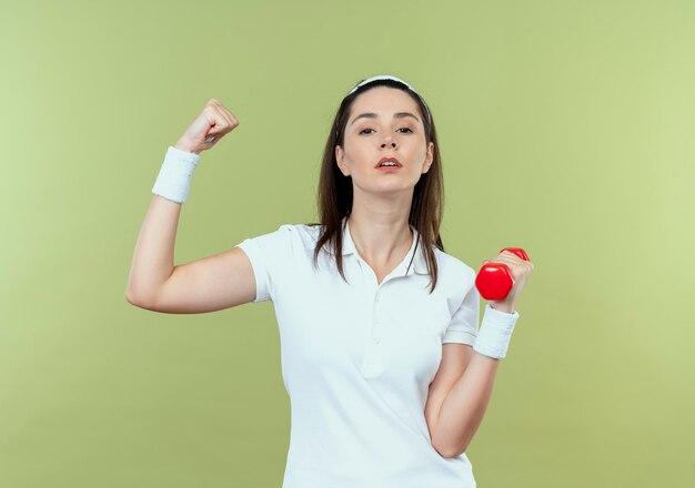 Young fitness woman in headband working out with dumbbell showing biceps  with serious face standing over light wall