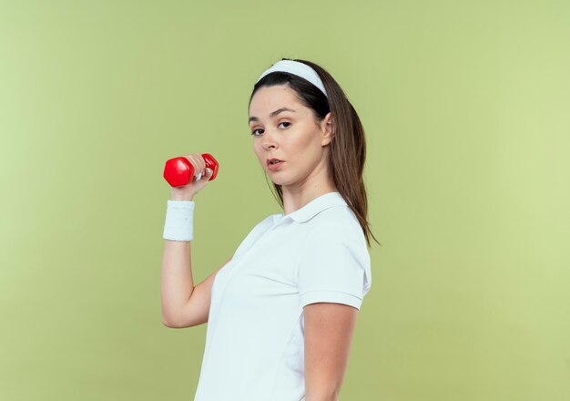 Молодая женщина фитнеса в оголовье, тренирующемся с гантелями с серьезным лицом, стоящим над светлой стеной