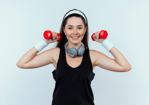 Young fitness woman in headband working out with dumbbells looking confident smiling standing over white wall