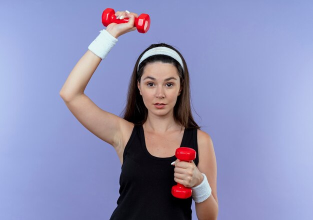 Young fitness woman in headband working out with dumbbells looking confident standing over blue wall