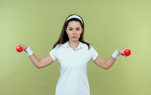 Young fitness woman in headband working out with dumbbells looking confident standing over light background