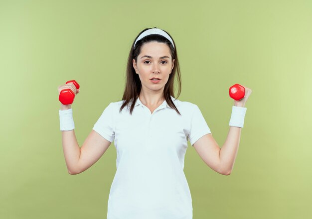 軽い壁の上に立って自信を持って見えるダンベルでワークアウトヘッドバンドの若いフィットネス女性