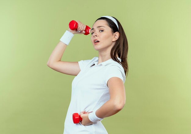 Young fitness woman in headband working out with dumbbells looking confident standing over light wall