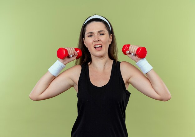 Young fitness woman in headband working out with dumbbells looking confused standing over light background