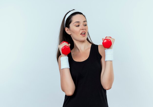 Young fitness woman in headband working out with dumbbells looking strained and confident standing over white wall