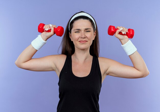 Молодая фитнес-женщина в головной повязке тренируется с напряженными и уверенными гантелями, стоя у синей стены