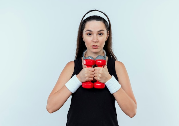 Giovane donna fitness in fascia lavorando con manubri con faccia seria in piedi sopra il muro bianco