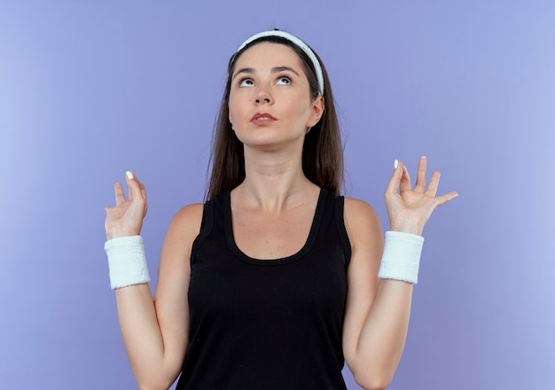 Бесплатное фото Молодая фитнес-женщина в повязке на голову, глядя вверх, делая жест медитации с пальцами, стоящими на синем фоне