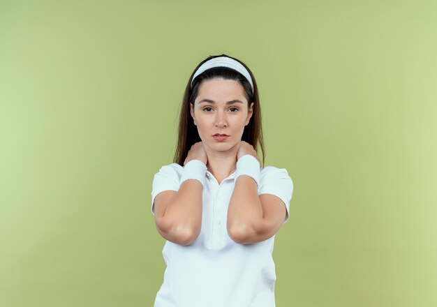 明るい背景の上に立っている深刻な顔でカメラを見て彼女の首に触れるヘッドバンドの若いフィットネス女性