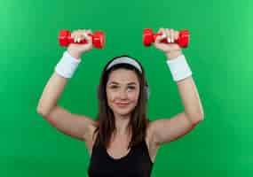 Бесплатное фото Молодая фитнес-женщина в головной повязке с наушниками, тренирующаяся с гантелями, уверенно улыбаясь, стоя на зеленом фоне