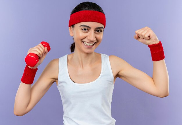 Бесплатное фото Молодая фитнес-женщина в спортивной одежде, держащая гантели, позирует и поднимает кулак, весело улыбаясь, стоя над фиолетовой стеной