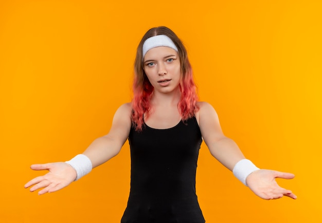 Бесплатное фото Молодая фитнес-женщина в спортивной одежде делает приветственный жест руками, стоящими над оранжевой стеной