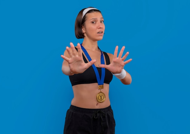 無料写真 彼女の首の周りに金メダルを持ったスポーツウェアの若いフィットネス女性は、青い壁の上に立って怖がっている手で防衛ジェスチャーをしています