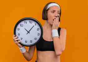 Бесплатное фото Молодая фитнес-женщина в спортивной одежде с повязкой на голову, держащая настенные часы, выглядит усталой и скучающей, зевая, стоя над оранжевой стеной