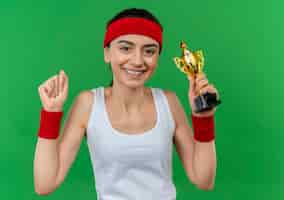 Бесплатное фото Молодая фитнес-женщина в спортивной одежде с повязкой на голову поднимает кулак, как победитель, уверенно улыбаясь, стоя над зеленой стеной