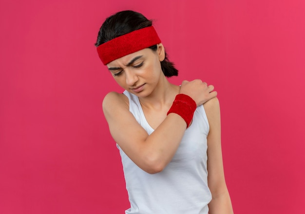 Бесплатное фото Молодая фитнес-женщина в спортивной одежде с повязкой на голову, касающаяся ее плеча, выглядит нездоровой, чувствуя боль, стоя над розовой стеной
