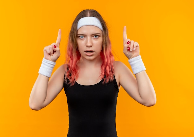 Бесплатное фото Молодая фитнес-женщина в спортивной одежде с удивленным указательным пальцем, стоящим над оранжевой стеной