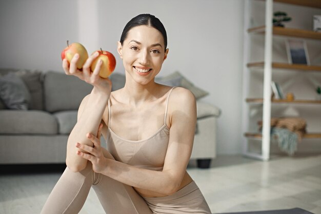 Young fitness woman sitting on yoga mat at home and holding fresh apples