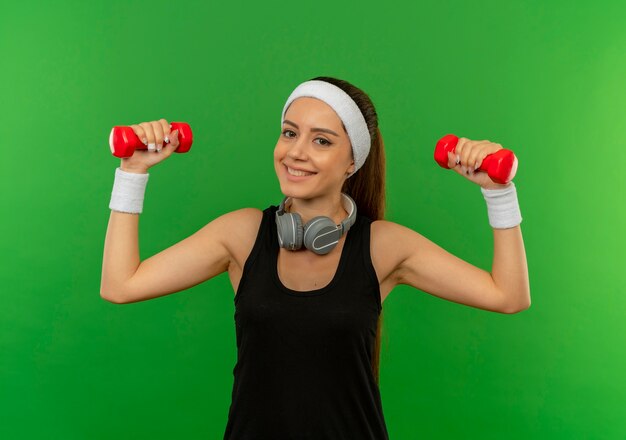 Молодая фитнес-женщина в спортивной одежде делает упражнения с двумя гантелями, весело улыбаясь и радостно улыбаясь, стоя над зеленой стеной