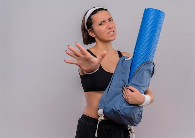 Giovane donna di forma fisica in abbigliamento sportivo che tiene zaino con materassino yoga facendo il fanale di arresto con la mano aperta con espressione disgustata in piedi sopra il muro bianco