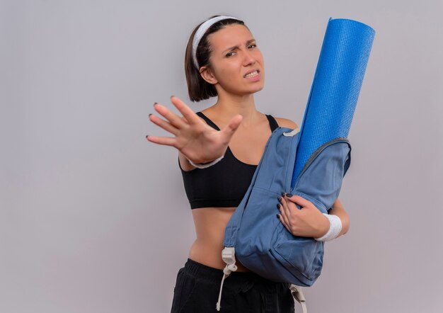 Молодая фитнес-женщина в спортивной одежде, держащая рюкзак с ковриком для йоги, делает знак остановки с открытой рукой с выражением отвращения, стоя над белой стеной