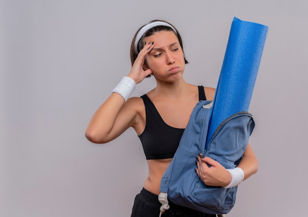 Молодая фитнес-женщина в спортивной одежде, держащая рюкзак с ковриком для йоги, глядя в сторону растерянной и усталой дующей щеки, стоя над белой стеной