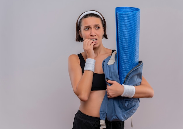 Young fitness woman in sportswear holding backpack with yoga mat looking aside stressed and nervous biting nails standing over white wall